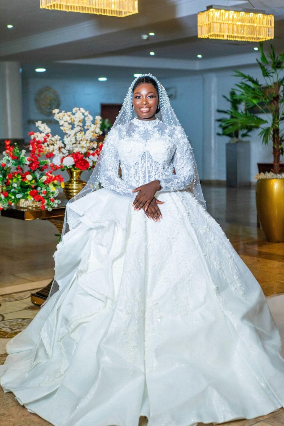 Beautiful bride in a luxurious white wedding dress with lace accents, standing in a decorated wedding venue with floral arrangements and modern lighting. Perfect for bridal fashion and wedding inspira.
